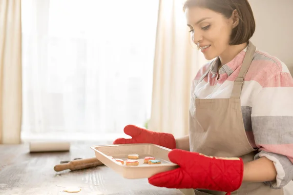 オーブンから取り出した自家製のクッキーでホットトレイを保持しているエプロンと手袋のかなり若い主婦 — ストック写真