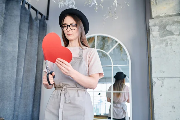 年轻的女人在准备情人节的时候 穿着围裙 戴着帽子 拿着大红纸的心和剪刀 — 图库照片