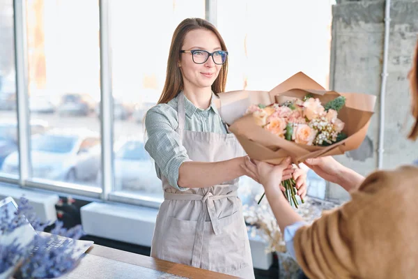 年轻的花商或店员在围裙和眼镜传递花束给买家或客户 — 图库照片