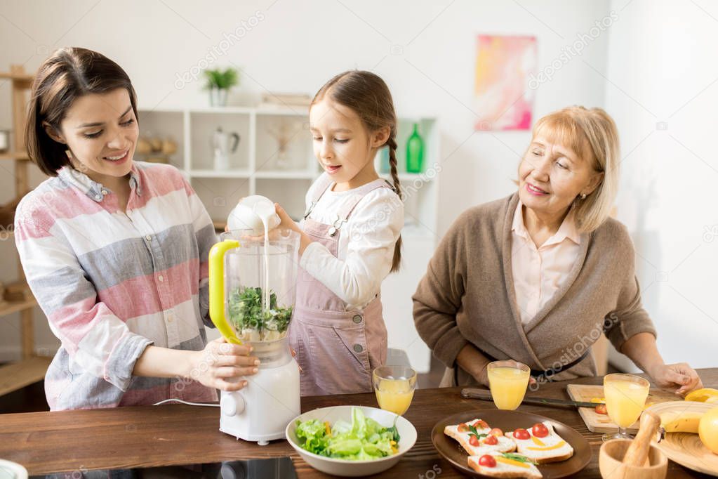 Adorale girl pouring fresh milk into electric blender while helping her mom with making smoothie