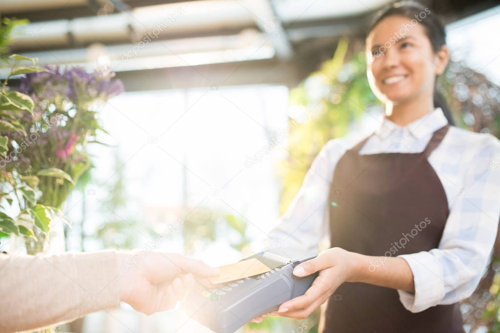 Young florist shop assistant holding payment machine while client or buyer paying for flowers