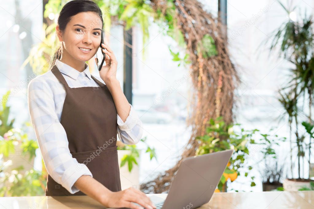 Young attractive cheerful woman in workwear using modern gadgets while working in florist shop