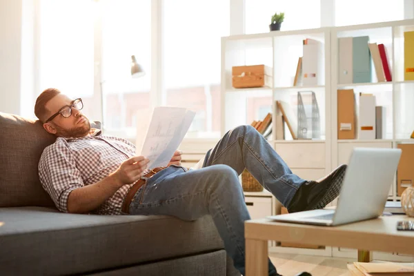 自宅で仕事中にラップトップの前でソファに横たわっているいくつかの論文と若いリラックス物思いビジネスマン — ストック写真