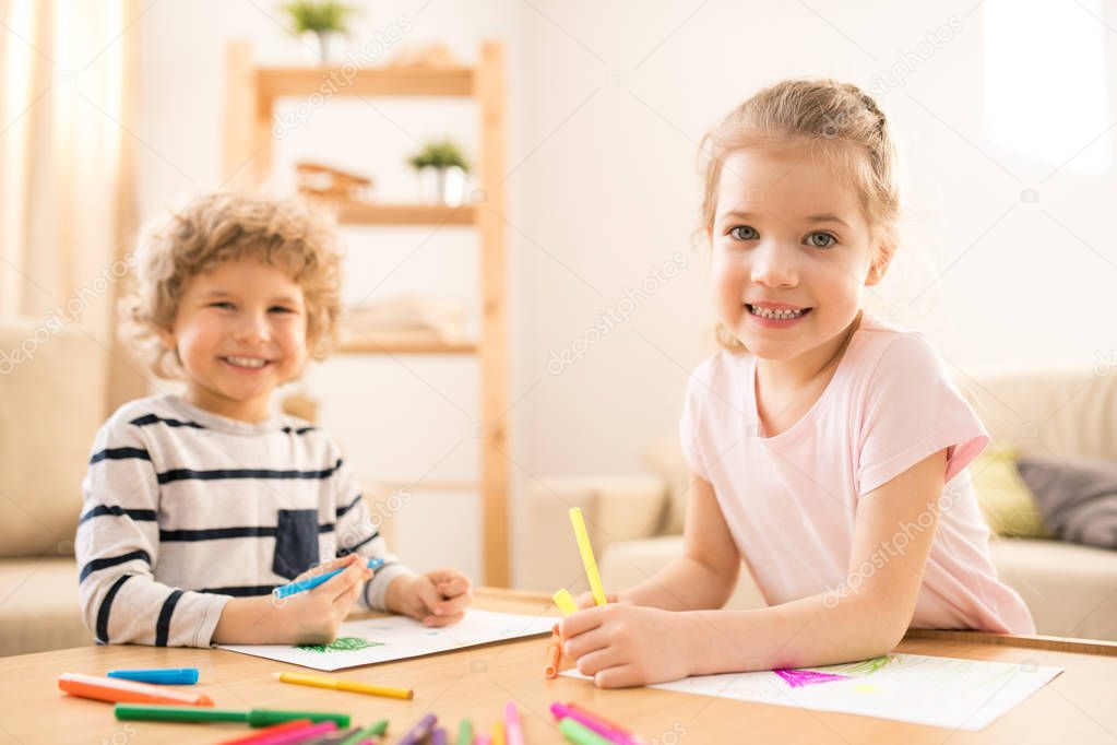 Happy little adorable girl and boy looking at you while drawing pictures with crayons by table