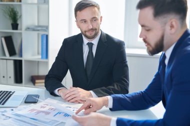 Takım elbiseli genç ve çekici bir işadamı ortak arıyor. Sözleşmeyi okuyor ve toplantıda metne işaret ediyor.