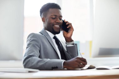 Mutlu başarılı genç ofis yöneticisi akıllı telefonla konuşurken işyerinde oturan ve defter notları yapma