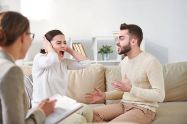 Genç rahatsız kadın bağırmak ve eller tarafından kulakları kapsayan onun sinir kocası aile oturumunda bir şey açıklamak için çalışırken