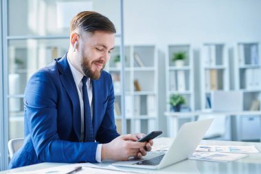 Ofiste işyerinde otururken akıllı telefonda çalma listesinde dolaşan kulaklıklı mutlu genç broker