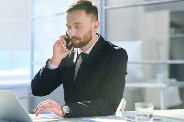 Dizüstü bilgisayarın önünde net gezinirken akıllı telefonda müşteri ile konuşurken formalwear ciddi genç komisyoncu