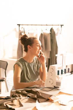 Yeni sipariş üzerinde çalışırken müşteriyle telefonda konuşan genç kadın terzilik dükkanında dikiş makinesi