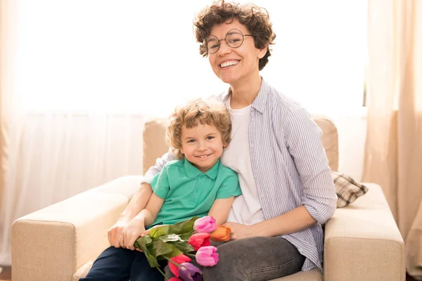 2人とも肘掛け椅子に座っている間に彼女の幼い息子を抱き 新鮮なチューリップの束を持つ明るい若い女性 — ストック写真