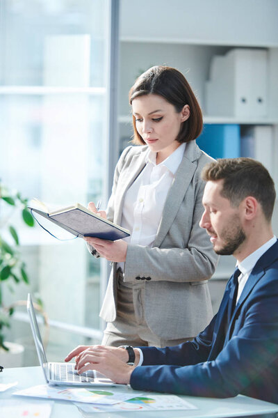 Young elegant businesswoman in suit reading her notes in notebook while her colleague entering or searching information in the net