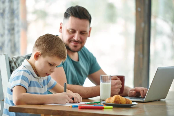 Little Son Ritning Med Hightändare Genom Bordet Medan Hans Pappa — Stockfoto