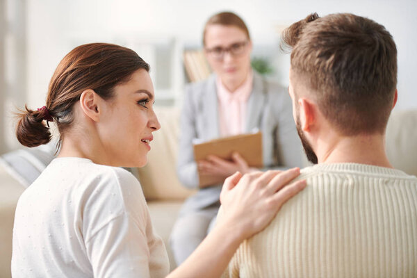 Young supportive wife looking at her husband while keeping hand on his shoulder during their visit to counselor office