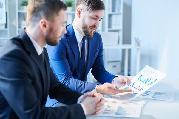 Один из молодых элегантных трейдеров показывает своим коллегам бумажные таблицы и диаграммы, объясняя ему данные
