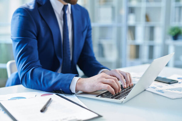 Elegant businessman in suit typing on keypad while sitting by desk in front of laptop in office