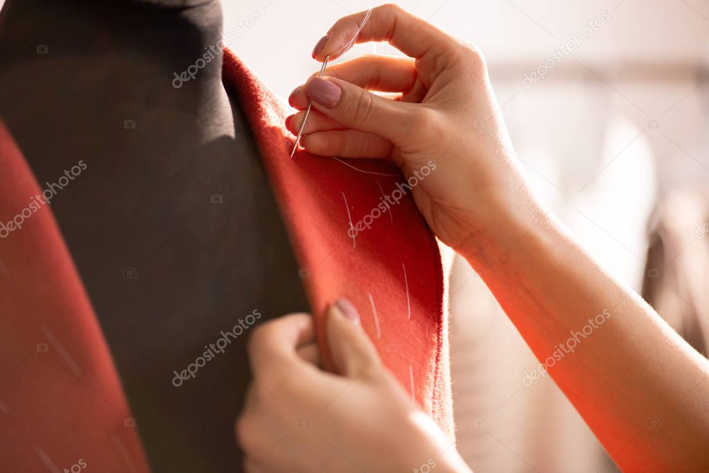 Hand of young female with needle and white thread working over collar of red coat on dummy