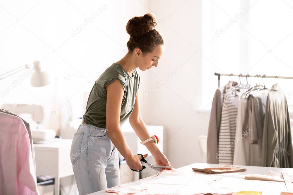 Young woman cutting fabric by edge of paper pattern while making new item for seasonal fashion collection