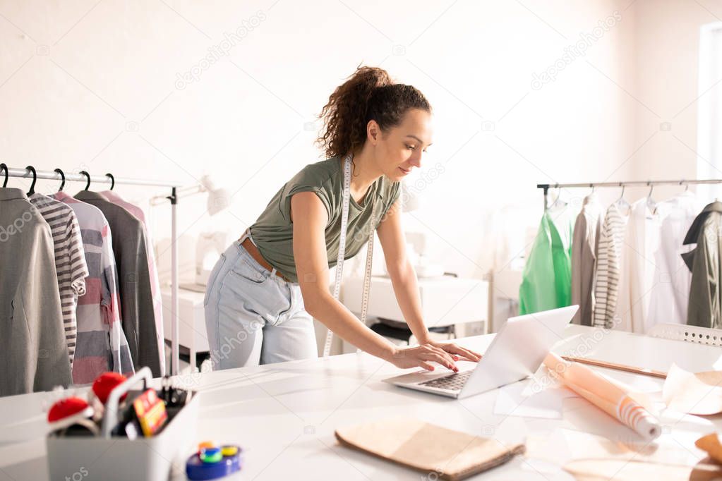 Young brunette fashion designer in casualwear bending over desk with laptop while surfing in the net for new creative ideas