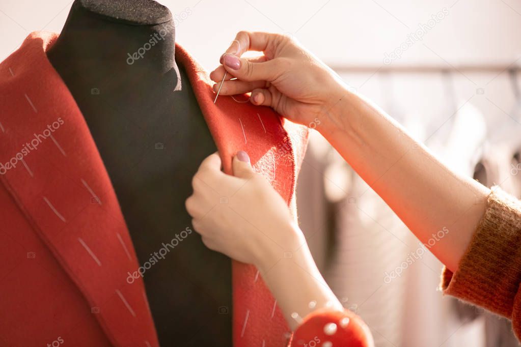 Hand of professional tailor using needle with white thread while sewing collar of red coat for one of clients