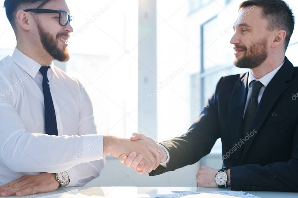 Two young successful business partners shaking hands over workplace and looking at each other with smiles