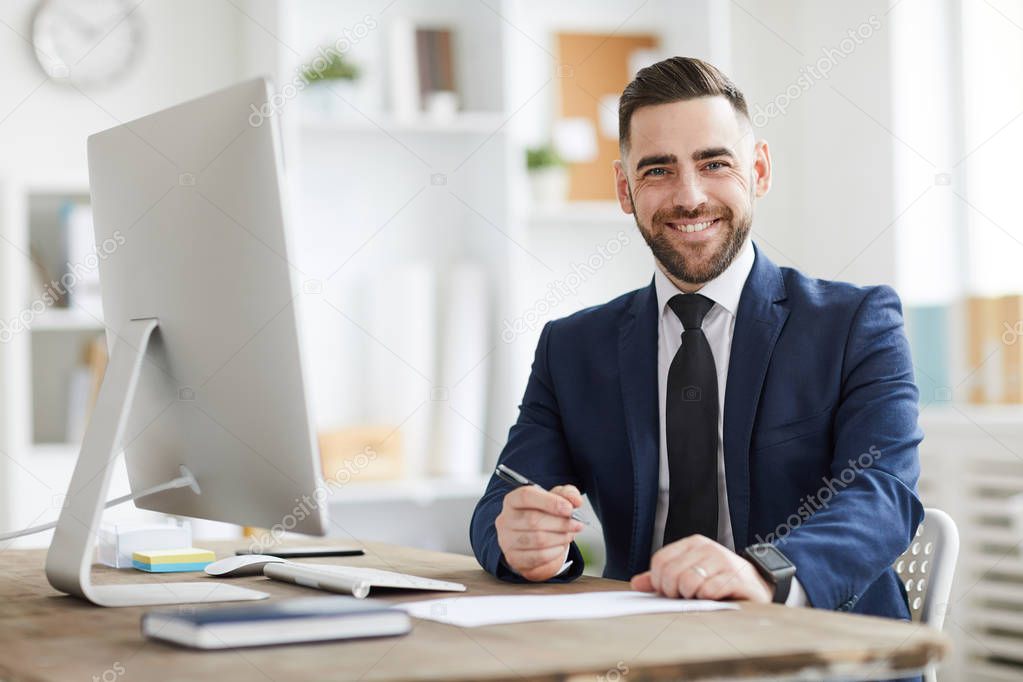 Young successful businessman in formalwear looking at you with smile while sitting by workplace in office
