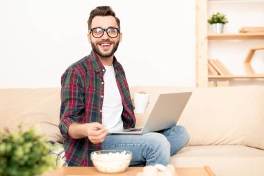 Genç gülen ev idarecisi gündelik kıyafetleriyle kanepede dinlenirken patlamış mısır ve çay içiyor.