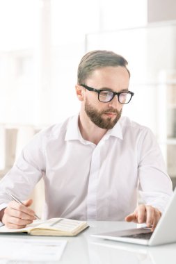 Young entrepreneur touching keys of laptop keypad while looking at display and making notes in notebook while working over project