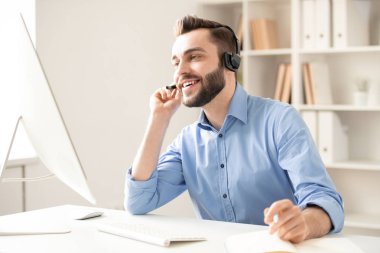 Young friendly operator looking at computer screen while talking to clients through video-chat in office