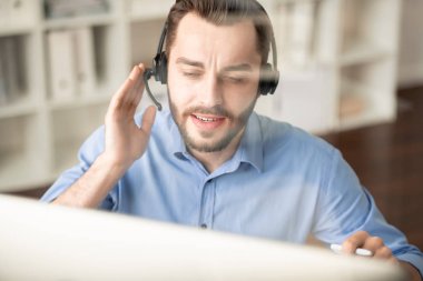 Young confident manager with headphone looking at computer screen while consulting clients and answering their questions