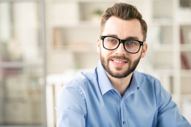 Young successful employee in eyeglasses and blue shirt sitting in office in front of camera by his workplace