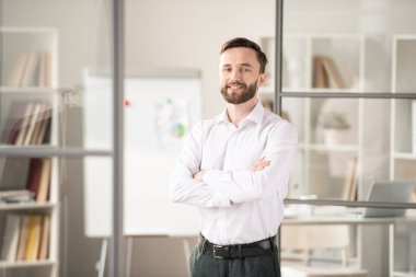Young bearded office worker crossing his arms on chest while standing in front of camera on background of workplace
