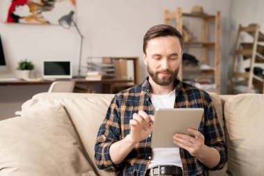 Young serious man with mobile gadget concentrating on network while looking at its screen and scrolling