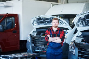Young successful cross-armed mechanic or repairman in overalls and flannel standing in workshop by car clipart