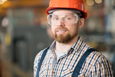Young successful bearded factory engineer in workwear, protective eyeglasses and helmet looking at you