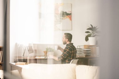 Young man with mug having drink while sitting by workplace in front of computer screen at home