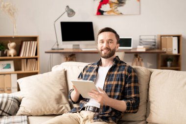 Young smiling employee with tablet sitting on couch at home in front of camera on background of workplace