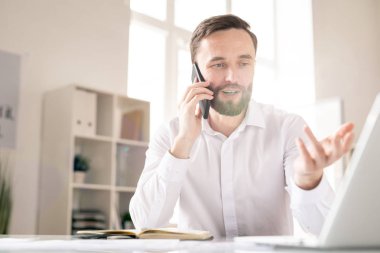Young man communicating to client or business partner on the phone while commenting online data on display of laptop