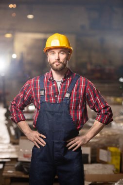 Young confident machinebuilder in workwear keeping his hands on waist while standing right in front of camera inside workshop