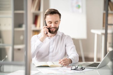 Sakallı genç çalışan, telefonda biriyle konuşurken not defterinde okuma veya not alma