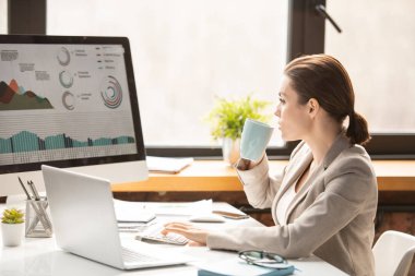İş yerinde çay ya da kahve içerken bilgisayar ekranındaki grafiksel verilere bakan genç bir finans analisti