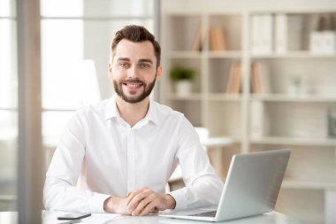 Young successful chief executive officer in white shirt sitting by desk in front of camera while planning work