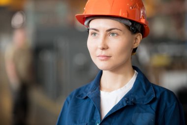 Young pretty female engineer in blue workwear and protective helmet looking after working process in factory