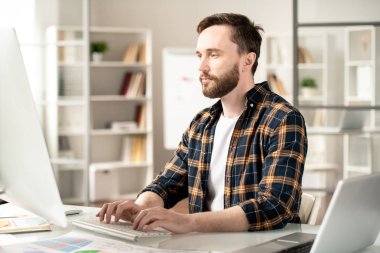 Young contemporary designer or programmer looking attentively at computer screen while working over new website