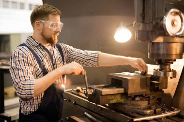 Young bearded technician in uniform and protective eyeglasses standing by industrial machine during working process