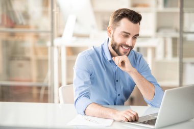 Mutlu genç ofis yöneticisi dizüstü ekran önünde oturan ve online video izlerken veya bilgi ile bakarak