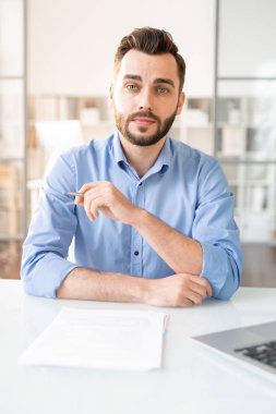 Young serious agent with pen sitting by desk with blank papers in front and listening to you