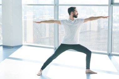 Young man in black leggins and white t-shirt stretching one leg while practicing yoga exercise with outstretched arms