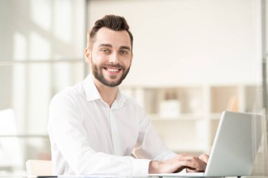 Young cheerful office manager with toothy smile surfing in the net or typing working plan while sitting in front of laptop