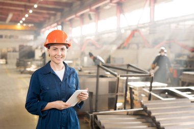 Young female engineer in helmet and blue overalls using modern gadget while working in factory